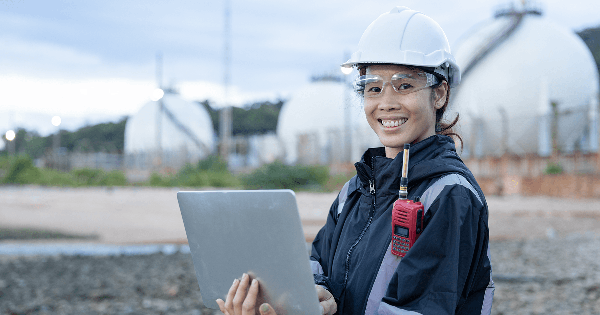 Field engineer wears a white helmet and dark jacket. They hold a laptop and have a hard hat on. The sky is grey. They stand in front of a work site.